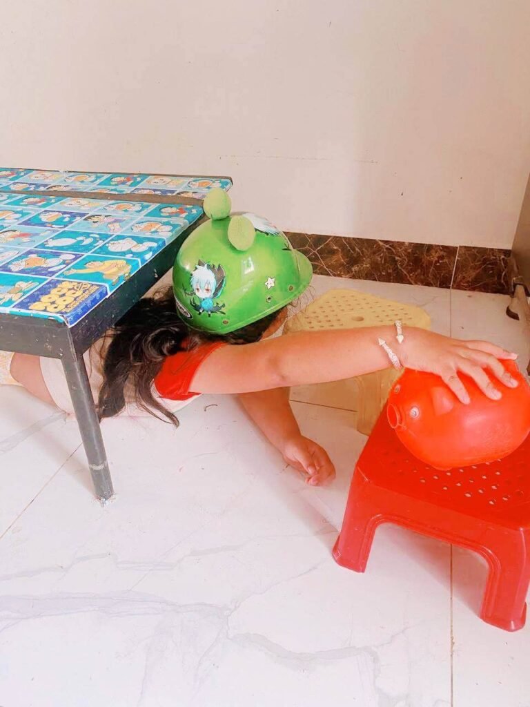 A young girl wearing a bright green bicycle helmet crawls under a low table on a tile floor, reaching out for an orange plastic piggy bank.