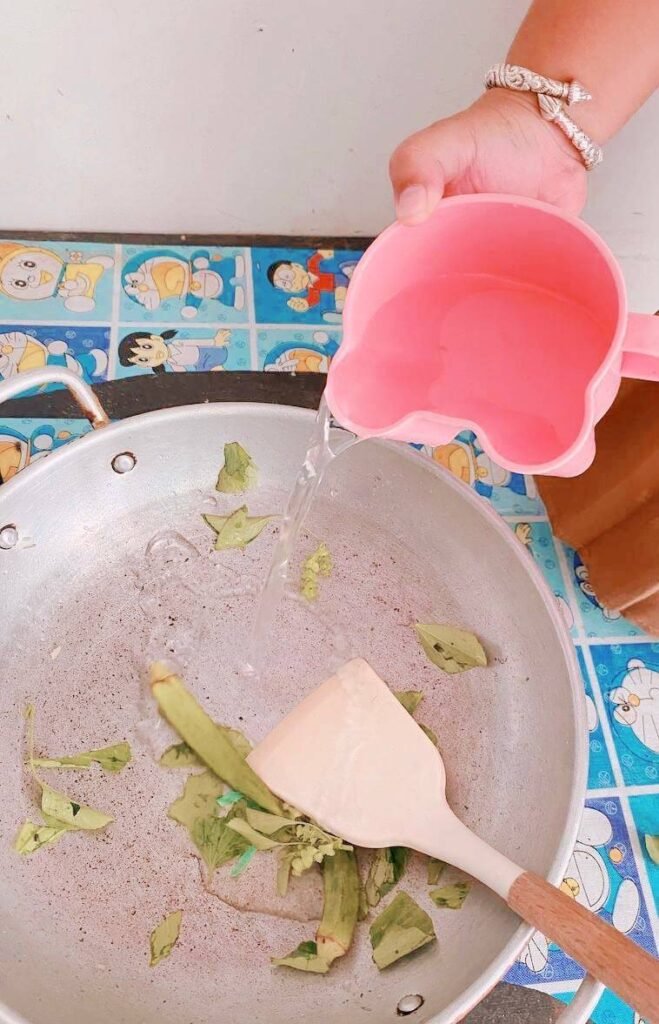 A child pouring water from a pink cup into a cooking pan with vegetables, completing the real-life math lesson.