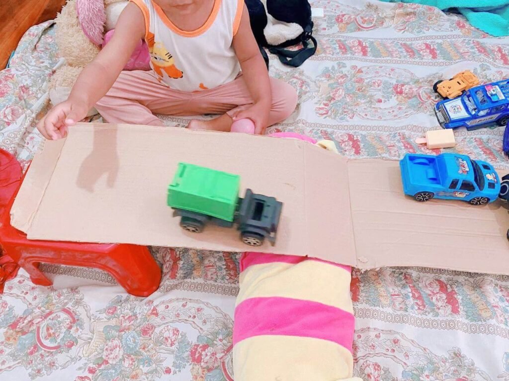 A toddler sits on a floral blanket and pushes a green toy dump truck down a single flat sheet of cardboard that is propped up on a small red plastic stool, creating a makeshift ramp. A blue truck waits at the top.