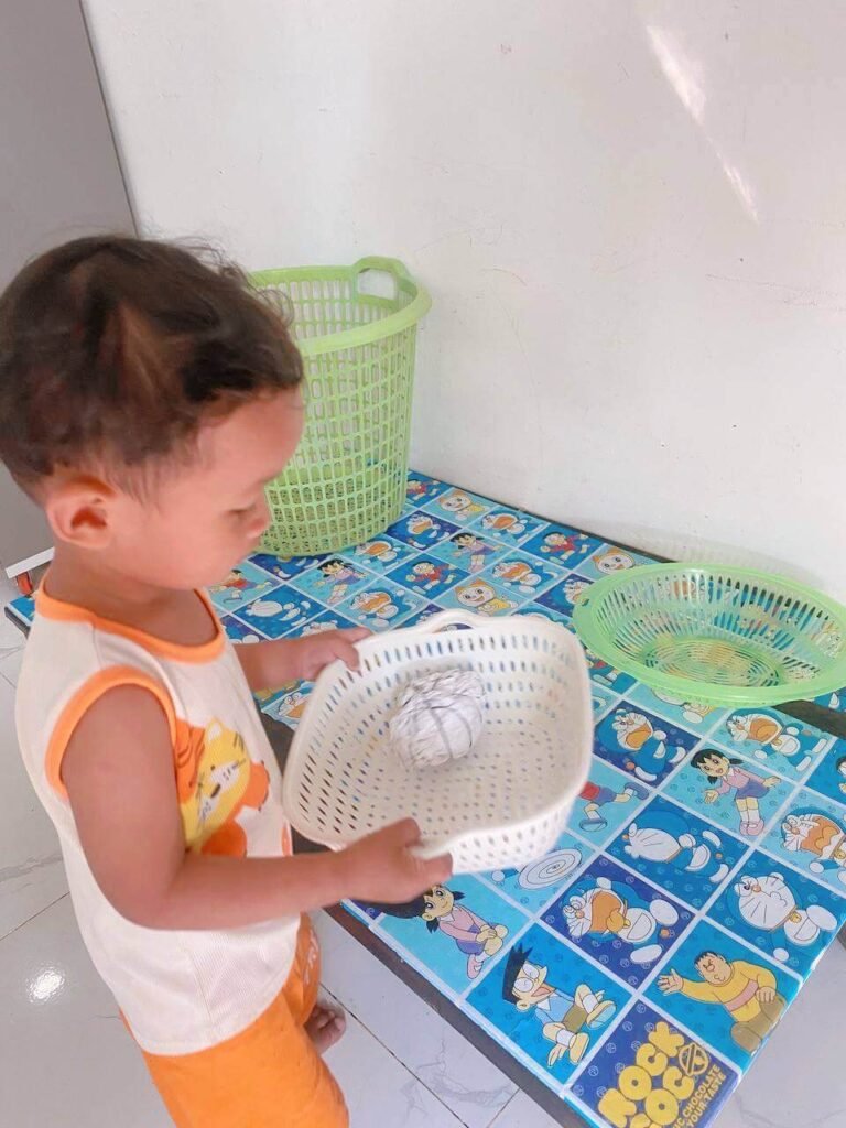 A toddler wears an orange tiger tank top and holds a white plastic strainer containing a homemade crumpled paper ball bound by thin hair rubber bands.