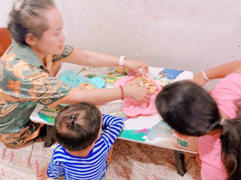 A mother leaning over a small table to serve mini pizzas on colorful plates to two young children.