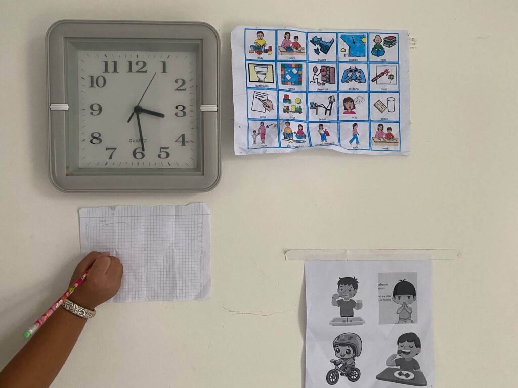 A white refrigerator door displaying an analog clock, a detailed visual schedule with behavioral icons, and a blank graph notepad with a pencil propped up.