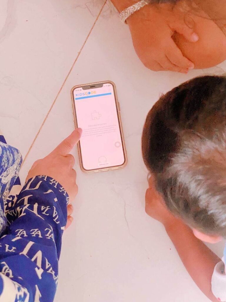 A parent's hand pointing to the Kids Code educational gaming app on an iPhone screen while a child looks closely to play together.