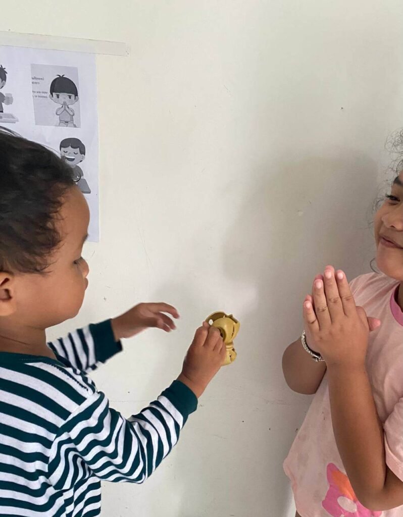 A young girl with her hands clasped smiling at her toddler brother during a family gratitude practice.