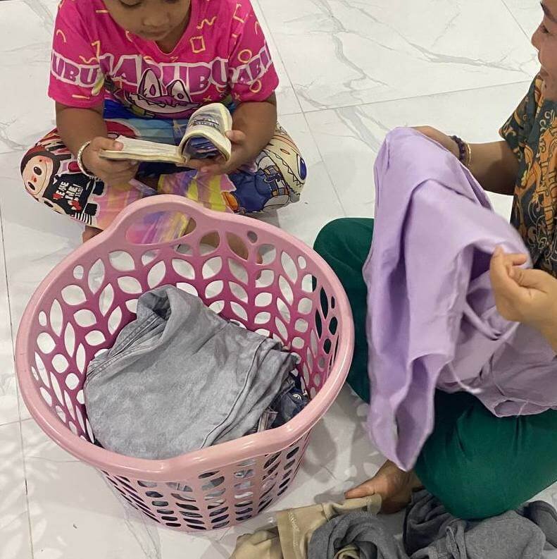 A parent folding purple and gray clothes from a pink laundry basket while a child sits nearby reading a book.