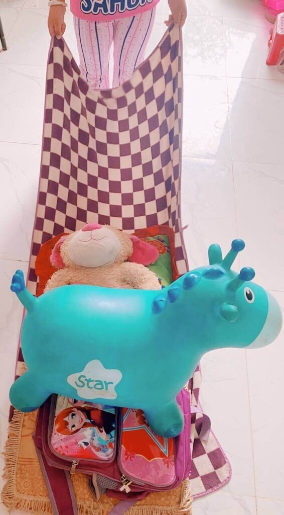 A child pulling a checkered blanket across the floor, loaded with a large blue toy, a stuffed animal, and a backpack as "cargo."