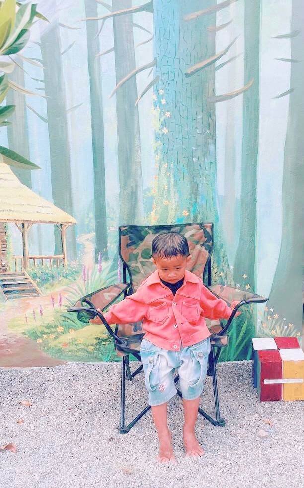 A young boy sitting quietly in a dark green camping chair outdoors, resting on a gravel surface in front of a painted forest mural.