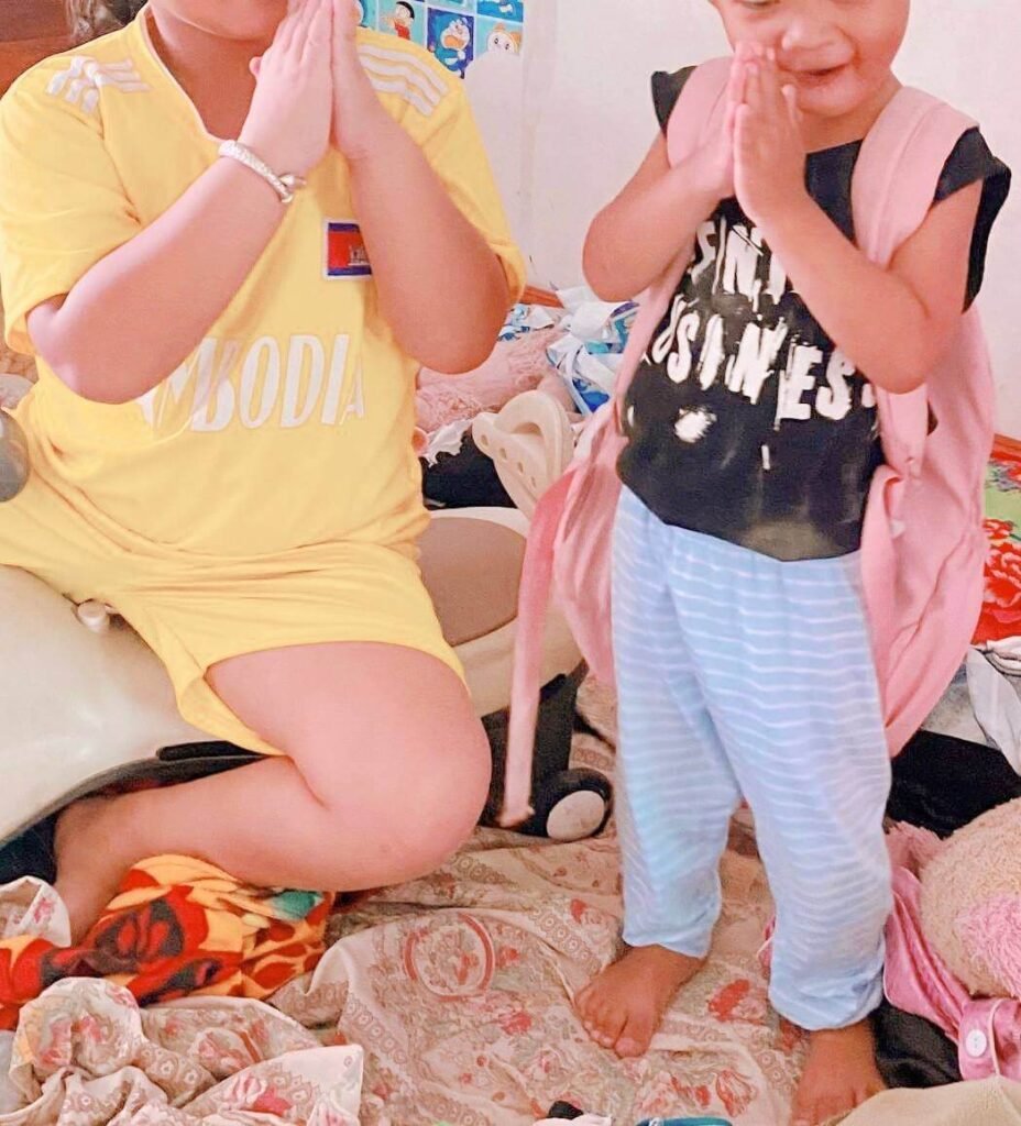 An older child wearing a yellow Cambodia shirt showing a toddler how to do the traditional Khmer Sompeah greeting.