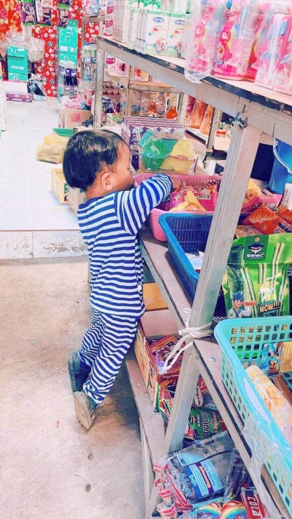 A young boy wearing blue and white striped pajamas reaching onto a lower shelf in a local market aisle.