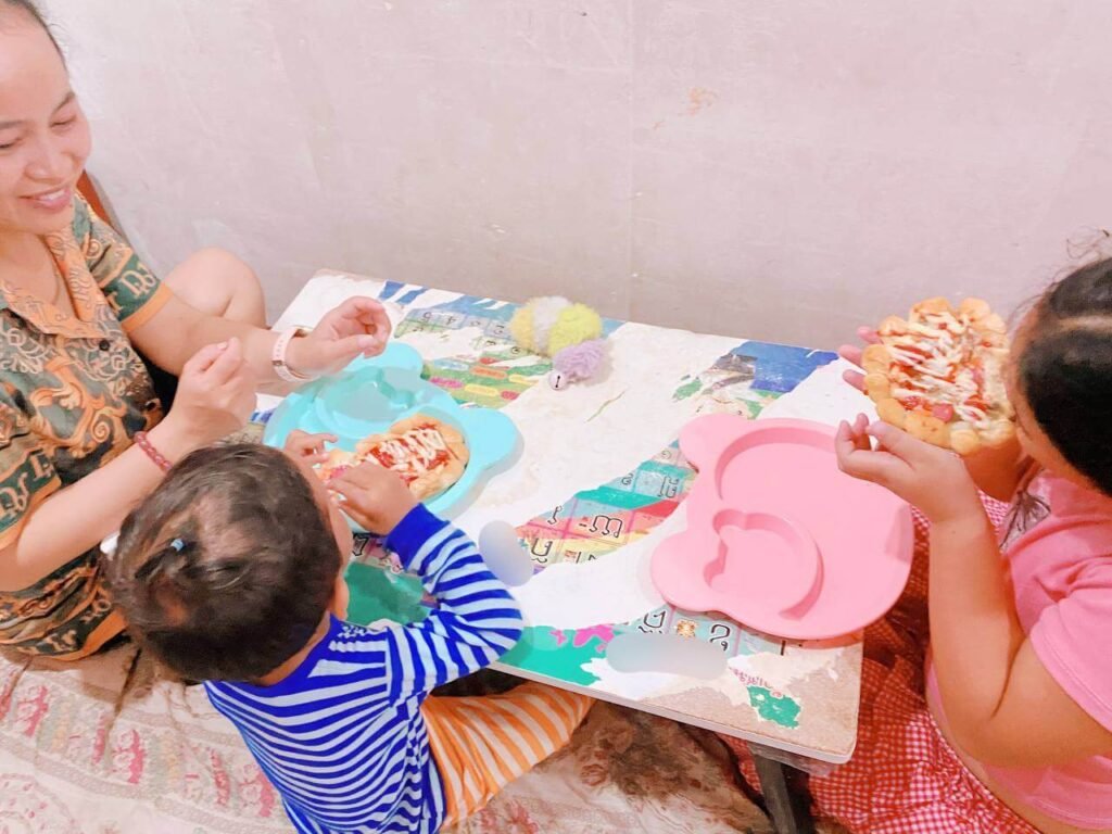A woman, a toddler in a striped shirt, and an older child in pink are sitting on the floor around a small table, happily holding and eating personal-sized pizzas.