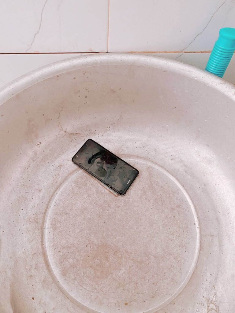 A black smartphone resting inside a large silver metal washbasin, simulating an electronics water damage accident for kids.