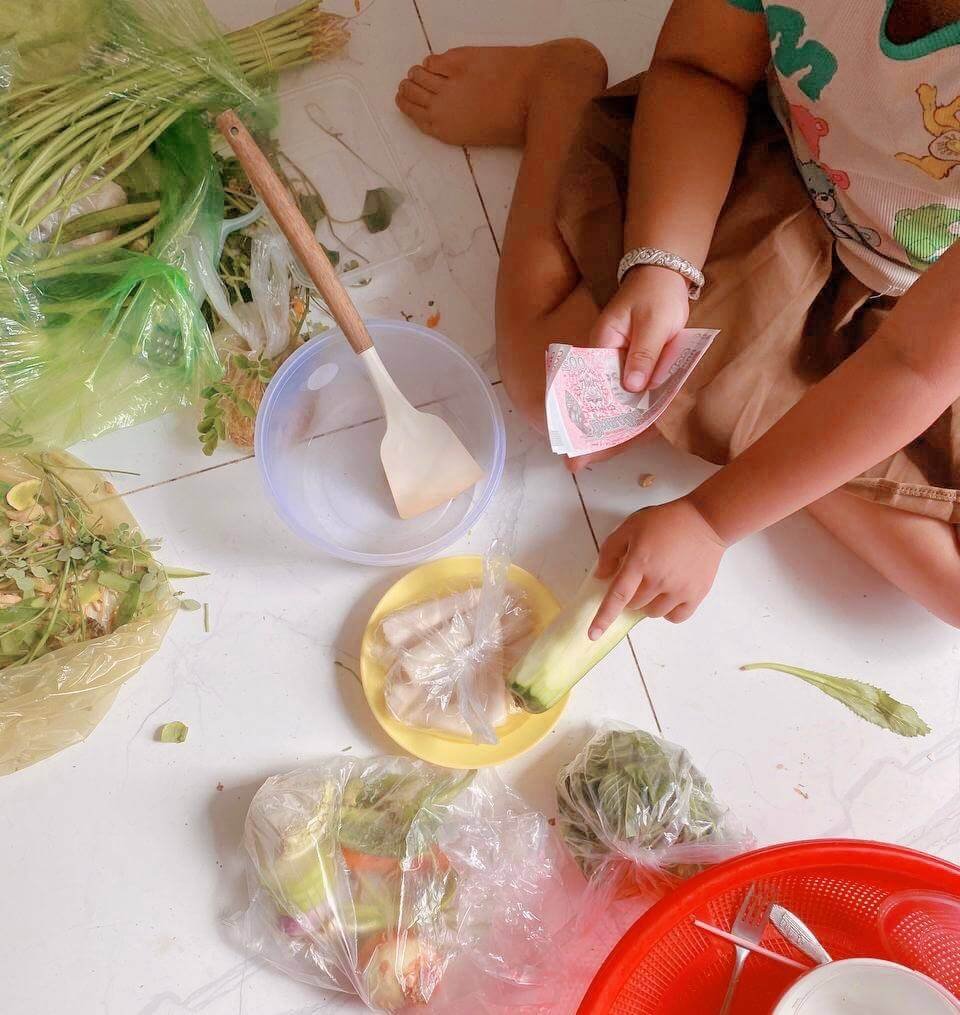 A child sitting on the kitchen floor holding Cambodian Riel notes, planning a dinner budget with fresh vegetables.