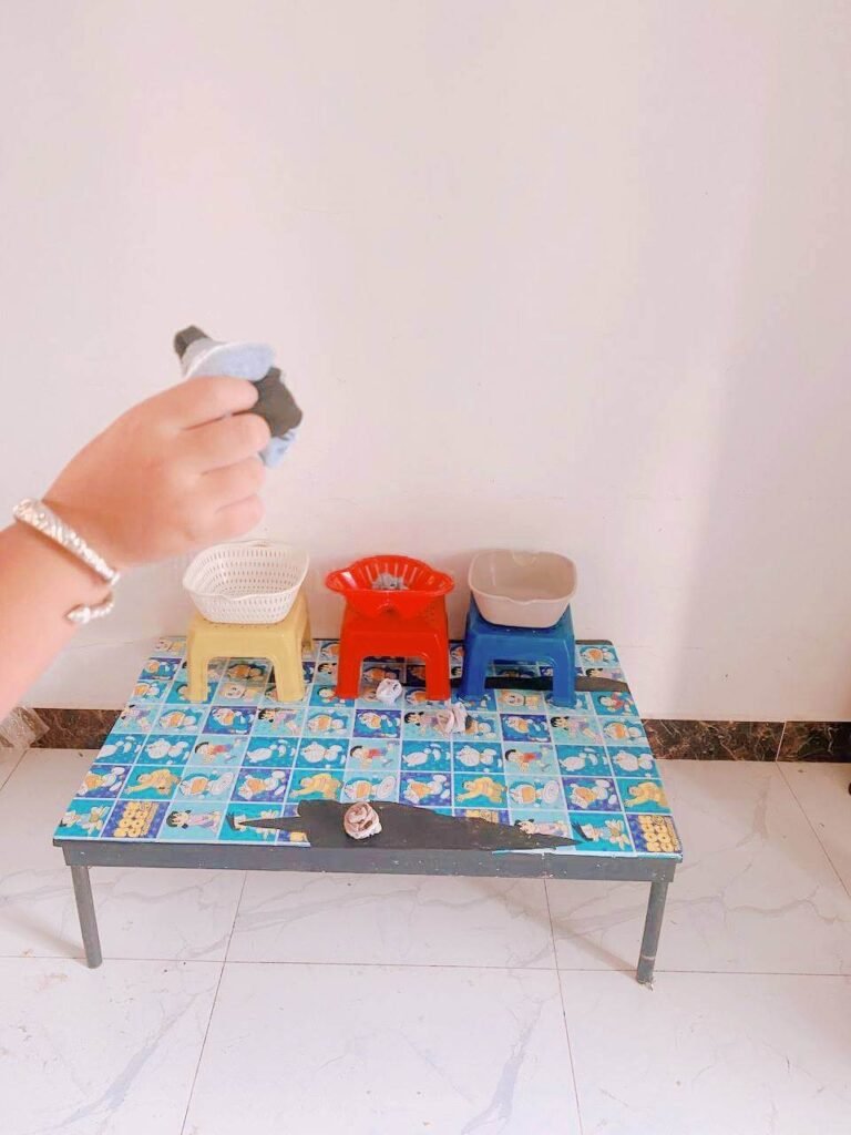 A hand holding a rolled-up pair of socks, aiming at three small, different-colored plastic baskets set up on a low table.