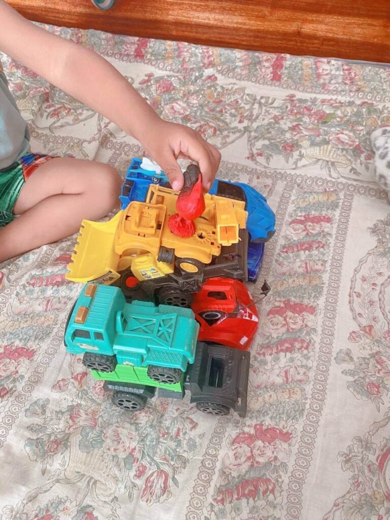 A small child, seen from the waist down, carefully places a small red plastic rooster figurine on top of a wobbly tower of yellow, blue, and green toy construction trucks stacked on a bed.