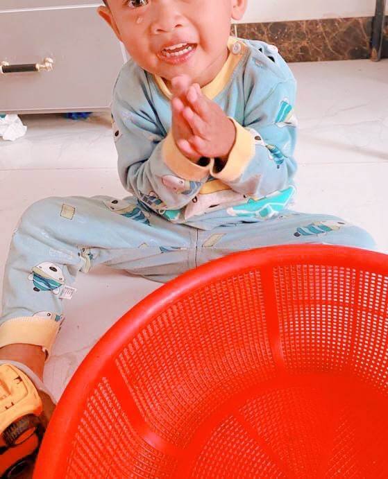 A toddler with a tear on his cheek sitting on the floor beside a red basket, holding his hands together to communicate.