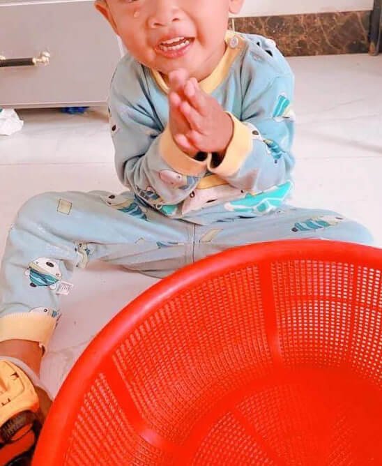 A crying toddler sitting on the floor with his hands clasped together, positioned next to an empty red plastic basket.