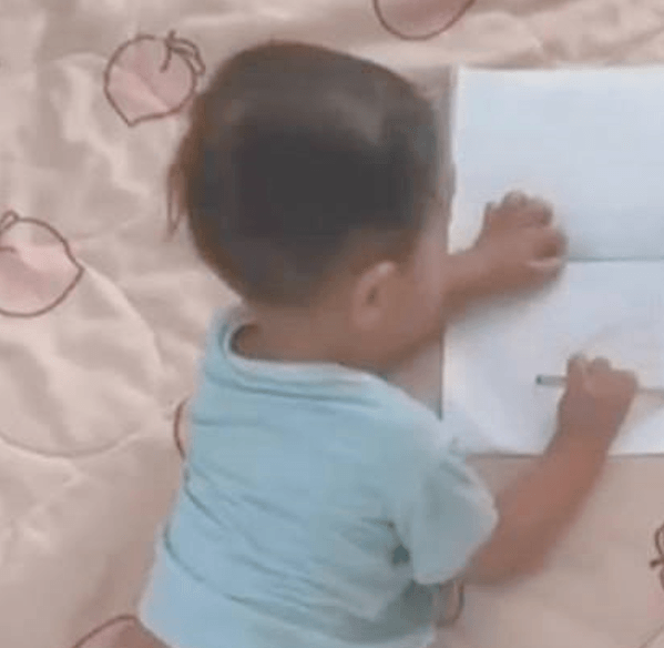 A young child lies on their stomach on a peach-patterned blanket, focused on drawing in a blank notebook with a pencil.