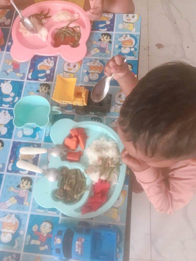 A young boy feeding himself rice and vegetables from a blue plate at a colorful dinner table.