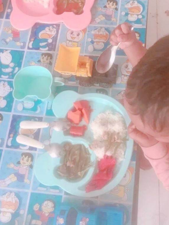 A toddler holding a metal spoon to eat from a light blue divided plate, with a yellow toy dump truck on the table nearby.