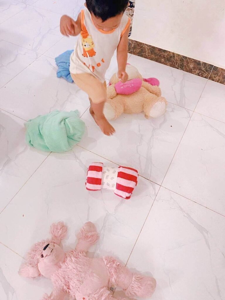 A toddler steps over a rolled-up red and white towel and stuffed animals placed on a white tile floor to create a makeshift walking path.