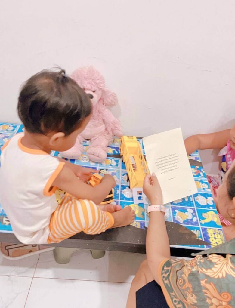 A toddler plays with a toy truck at a small table while a family member reads from a meeting agenda.