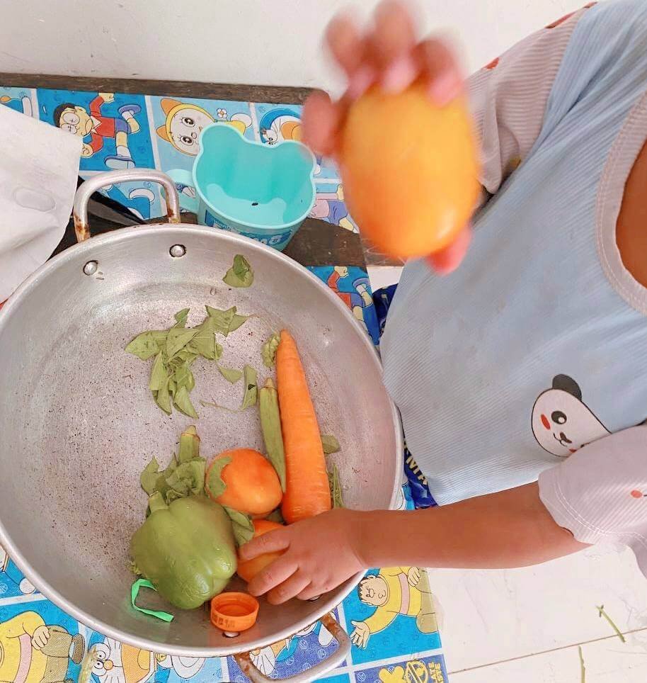 A small child in a blue panda shirt manipulates a green bell pepper, a whole carrot, and tomatoes inside a large metal wok. They hold another round orange object. Real vegetable scraps are seen on the floor below.