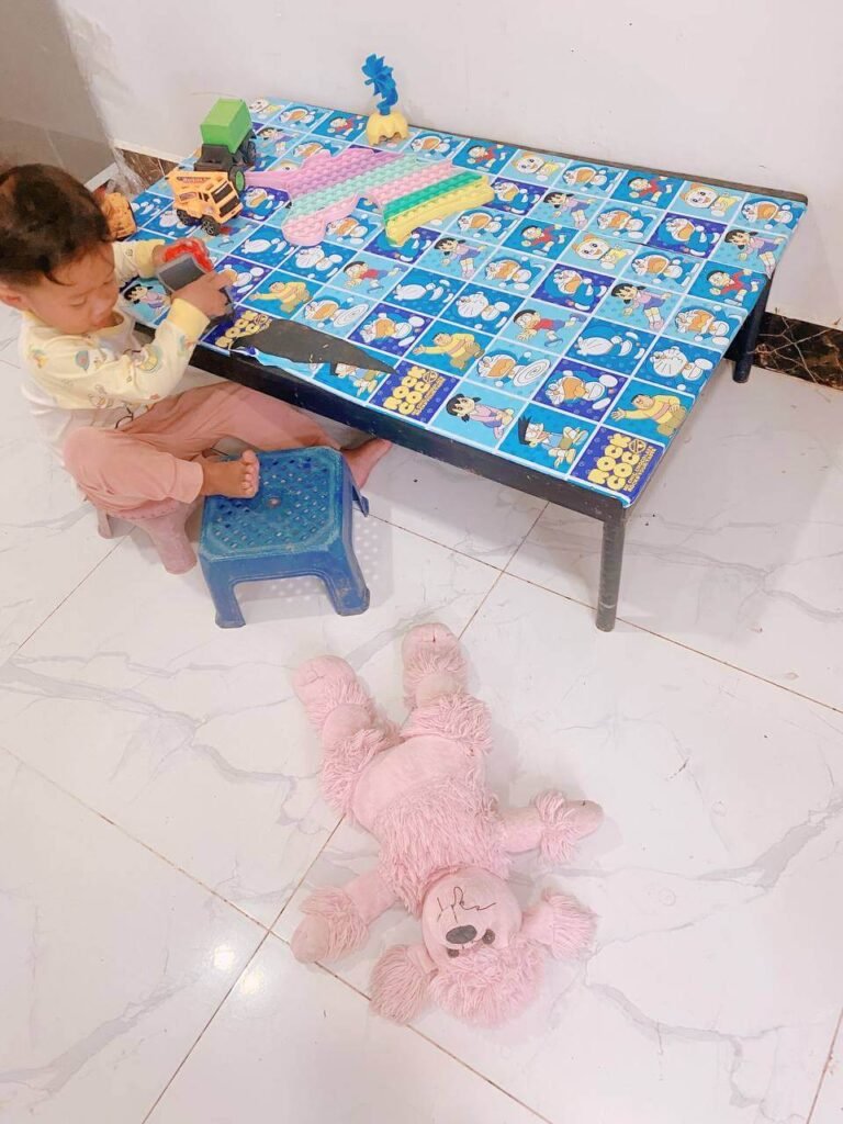 A toddler sits at a low play table with toy cars and a pop-it toy. On the white tile floor in the foreground lies a pink stuffed poodle, waiting to be rescued.