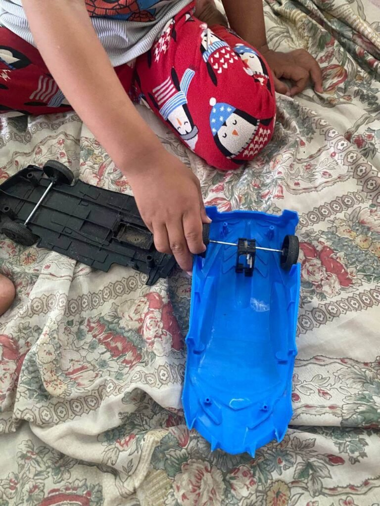 A child's hands carefully separate the blue plastic shell of a toy car from its black wheeled chassis on a floral bedspread.