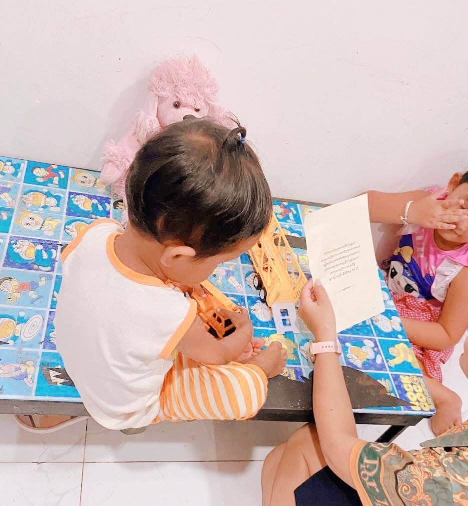 A toddler boy playing with a yellow toy crane at a blue table while his family has an everyday conversation.