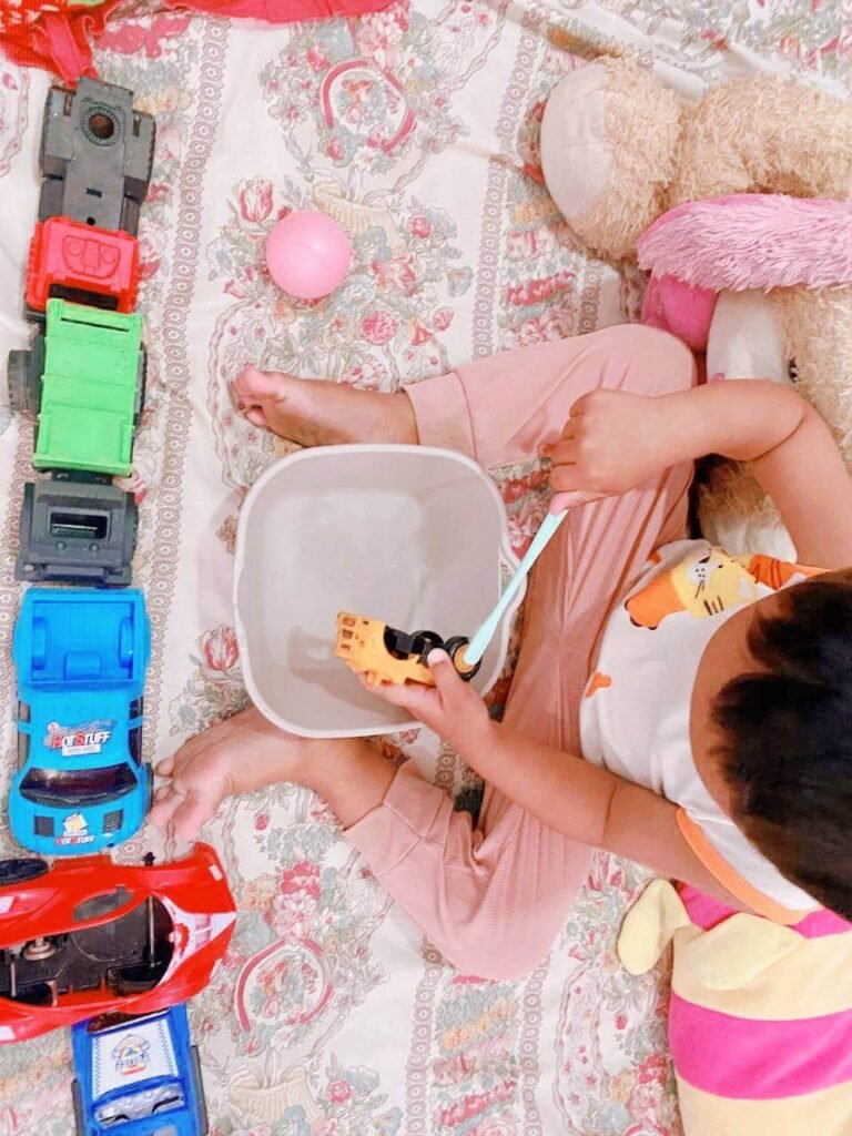 A toddler sits on a bed with a long line of toy trucks next to them, using a light blue toothbrush to scrub a yellow toy truck over a small plastic tub of water.