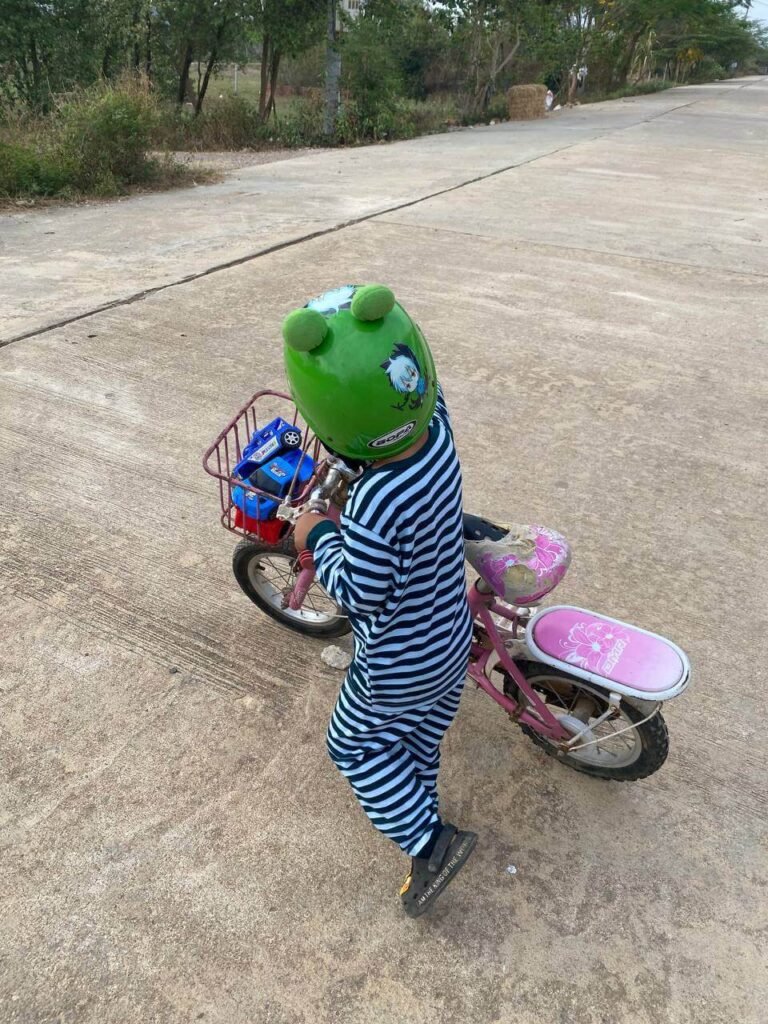 A small boy wearing a large green frog helmet standing next to an older sibling's pink bicycle on a paved road.
