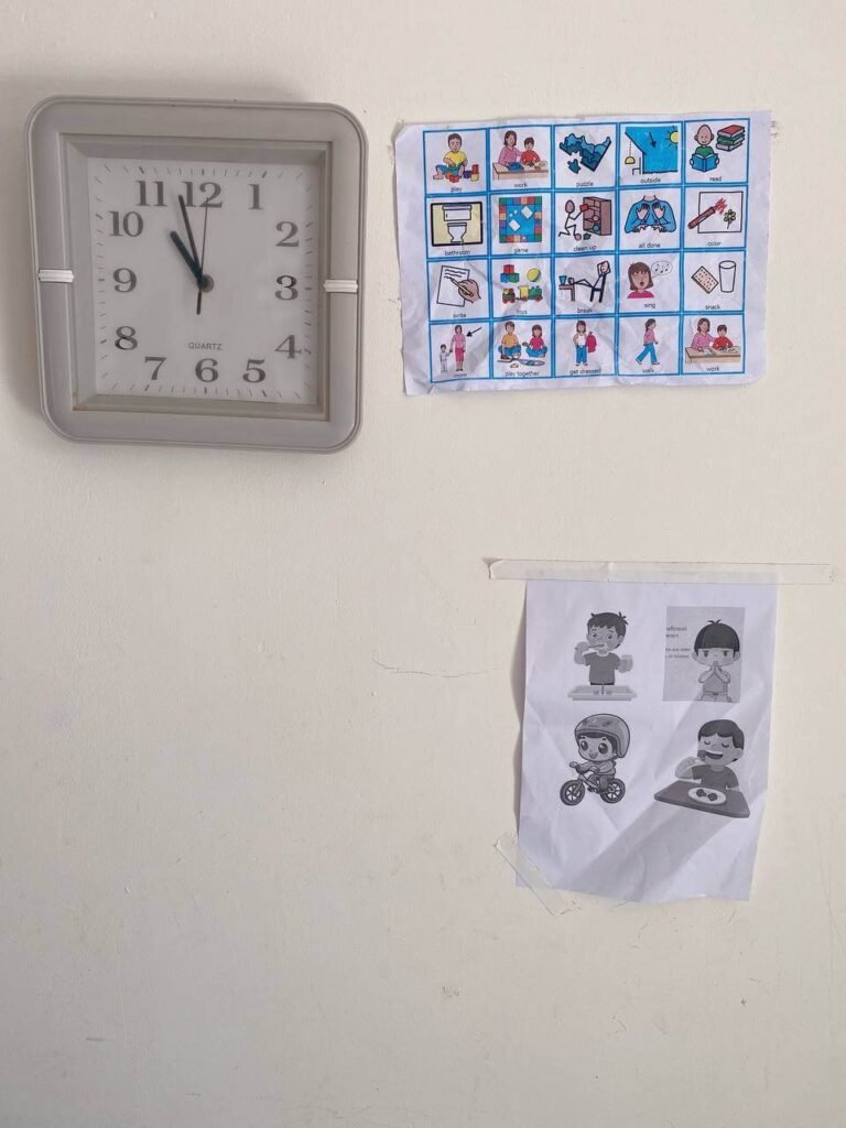A printed visual daily schedule chart with colorful activity pictures hanging on a wall next to a square grey wall clock.