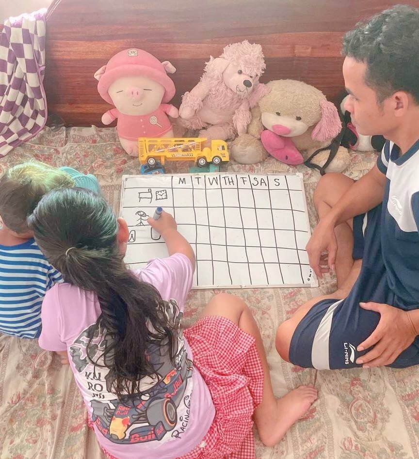 A young girl with long dark hair using a blue dry-erase marker to draw custom task icons on a whiteboard calendar grid.