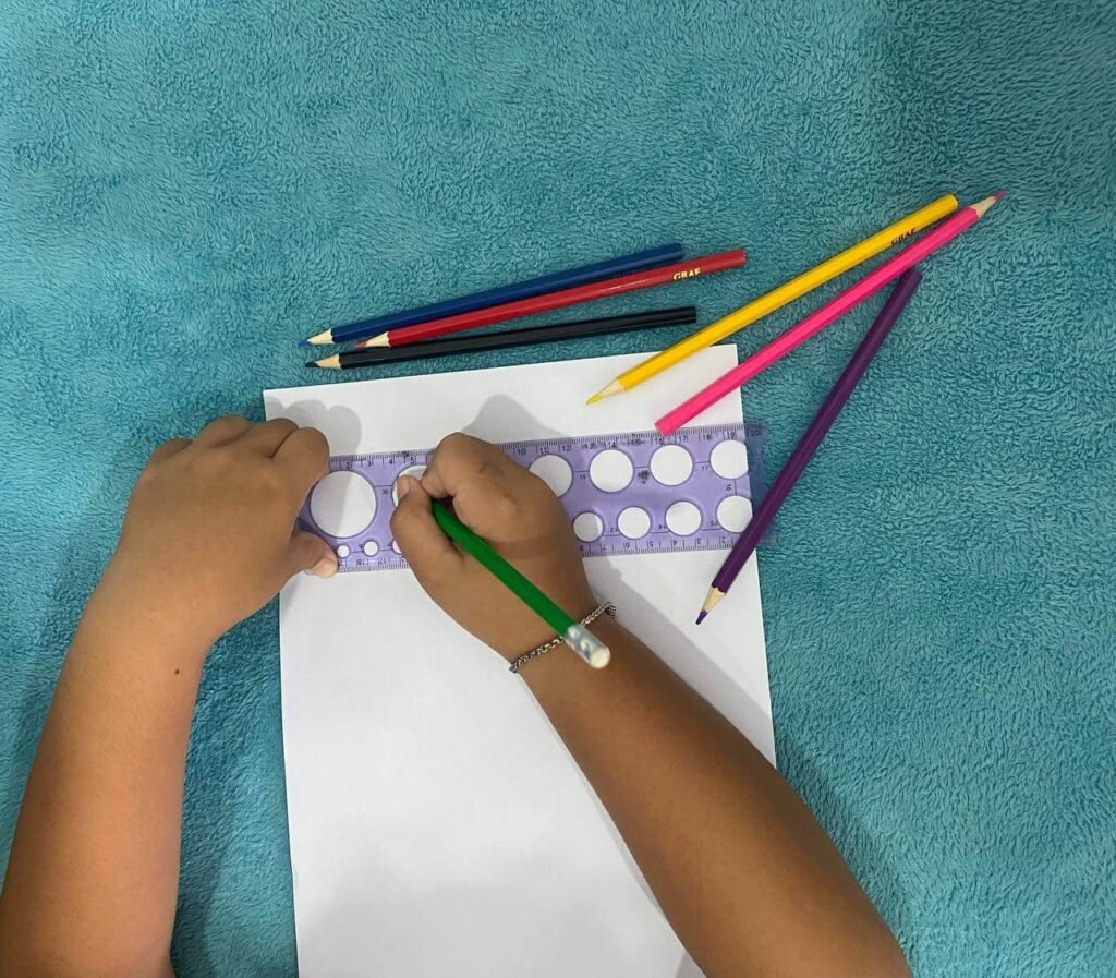A child's hands using a purple circle stencil ruler to start a dog drawing on blank white paper alongside colored pencils.