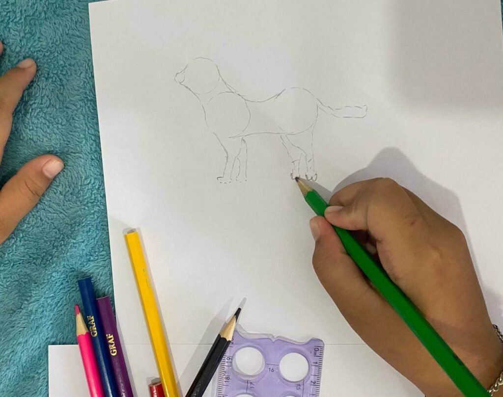 A child's hand using quick pencil strokes to draw paws and add zig-zag fur texture along the edges of a dog sketch.