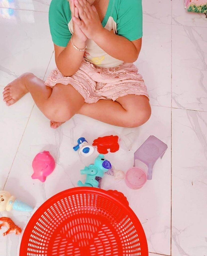 A young girl sitting on the floor making a Sampeah gesture with her hands, sitting next to scattered plastic toys and an empty red basket.