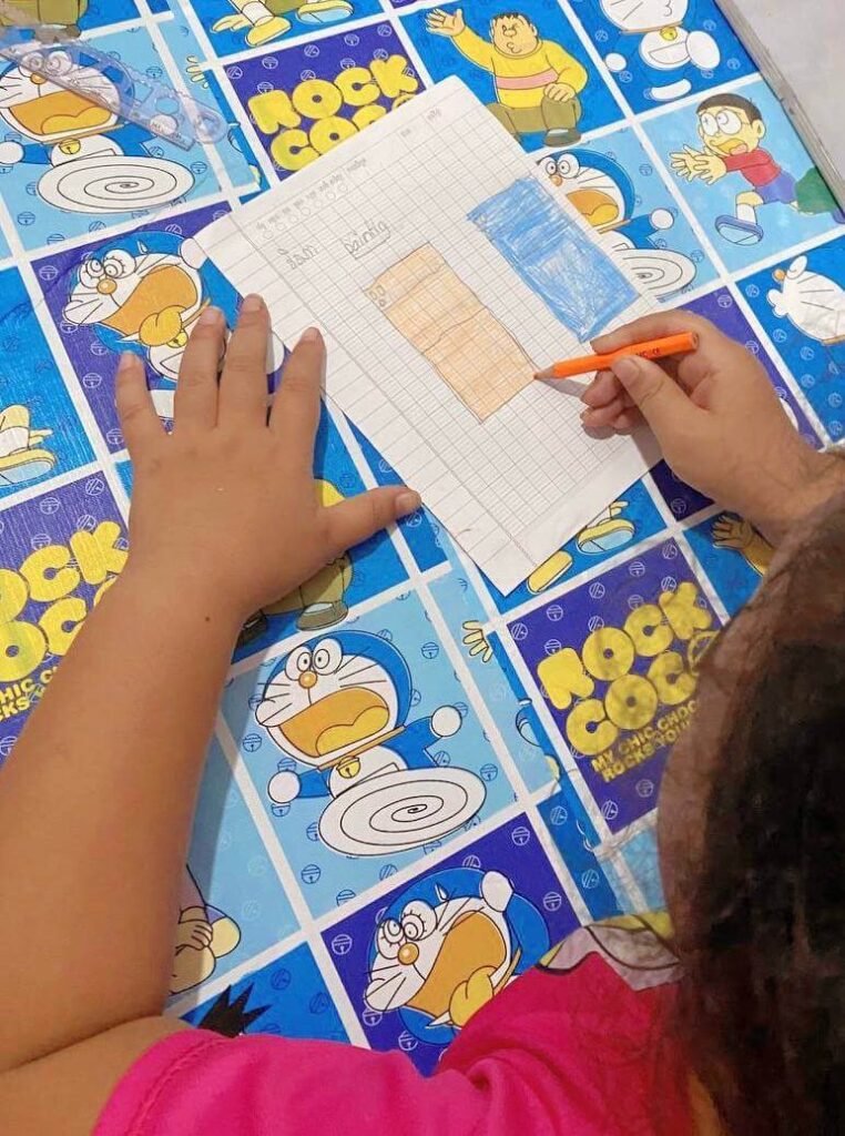 A top-down view of a child using an orange pencil to draw an orange rectangle and a teal rectangle on lined paper.