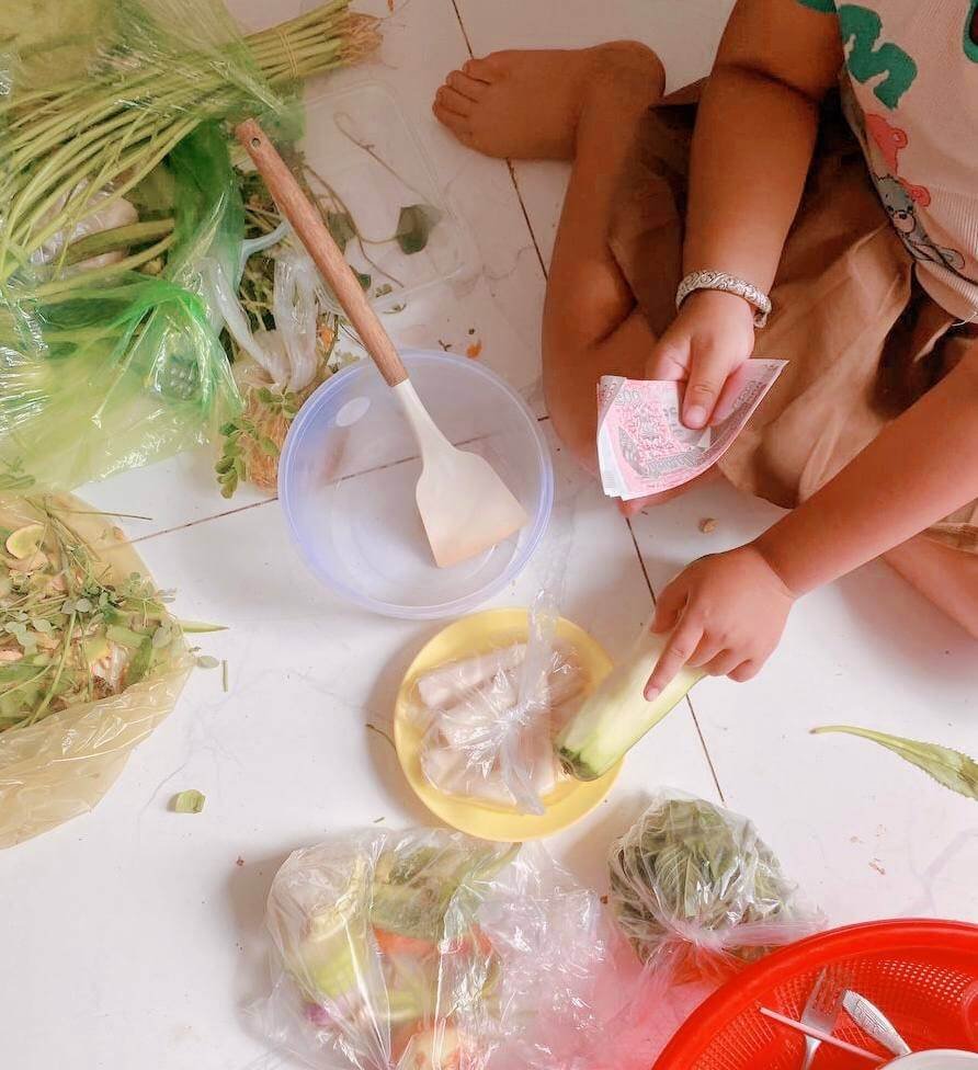 A child sitting on a tiled floor holding Cambodian Riel banknotes, surrounded by fresh vegetables and ingredients for Machur soup.