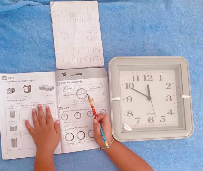 A child's hands pointing with a pencil at a time-telling exercise in a Khmer school workbook with a real square clock sitting next to it.