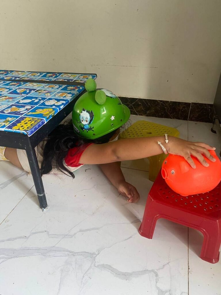 A child wearing a green toy helmet crawling under a small table to reach an orange plastic piggy bank sitting on a red stool.