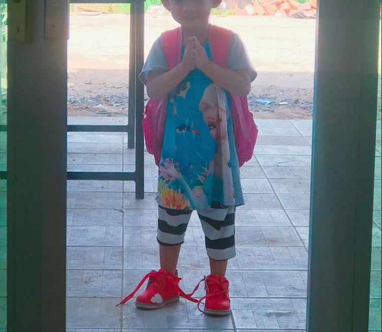 A young girl wearing a pink backpack and red shoes pressing her hands together to perform a traditional Cambodian sampeah at a doorway.
