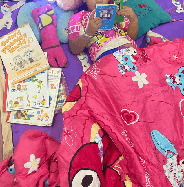 A young girl lying in bed under a pink blanket, reading a small book, surrounded by phonics workbooks and stuffed toys.