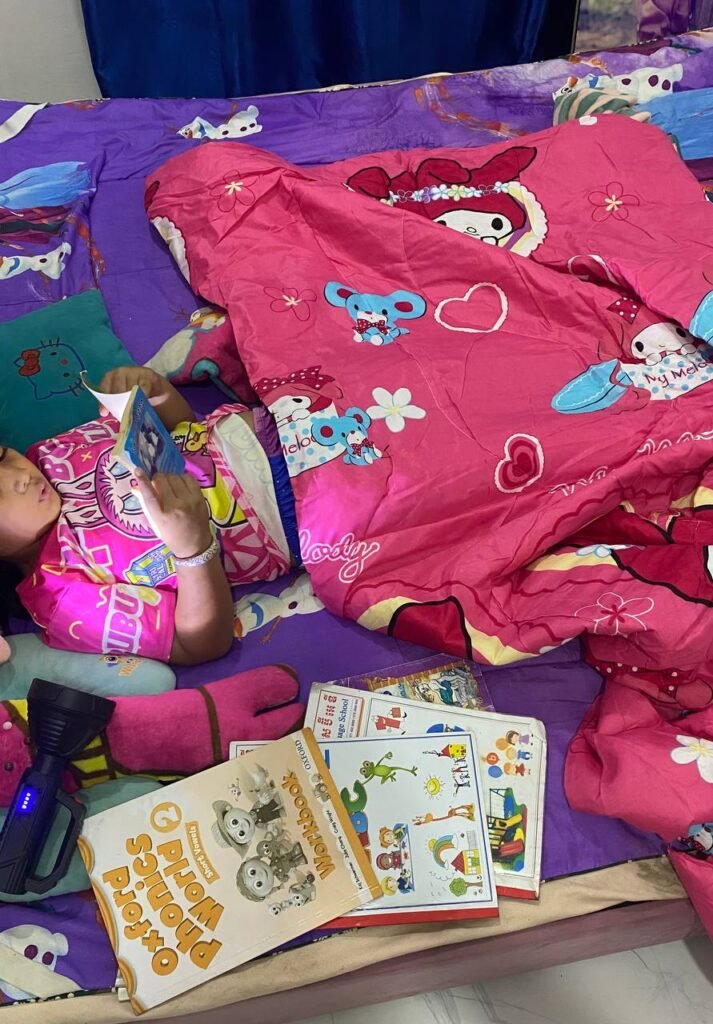 A young girl lying in bed under a pink blanket reading a small book, with an Oxford Phonics workbook lying next to her.