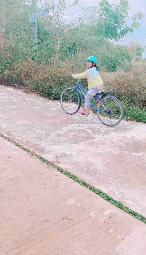 A young girl wearing a blue helmet and long clothes riding a blue bicycle on a quiet concrete road.