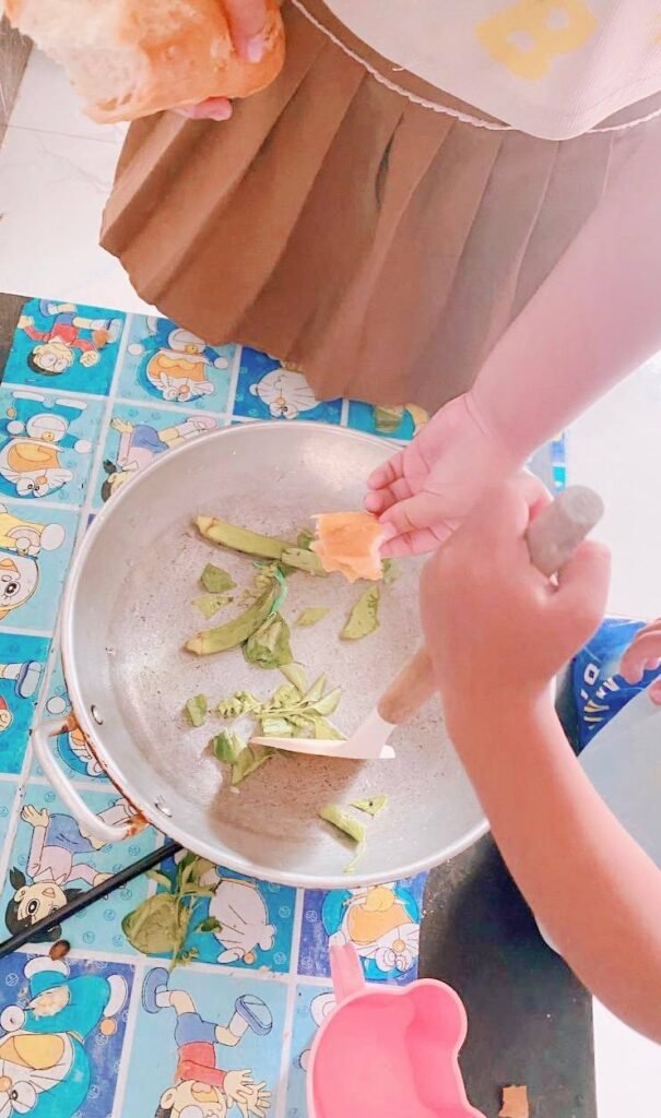 Two children standing at a table with a Doraemon tablecloth, testing a piece of bread in a pan with green vegetables.