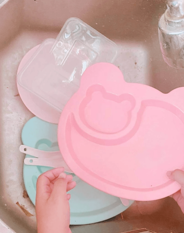 A child's hands holding a lightweight, pink bear-shaped plastic plate over a kitchen sink to practice dishwashing safely.