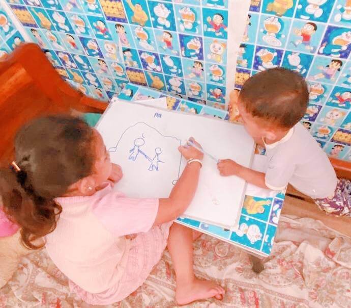 Two young children sitting at a small table, with the older girl drawing a stick-figure family on a small whiteboard.