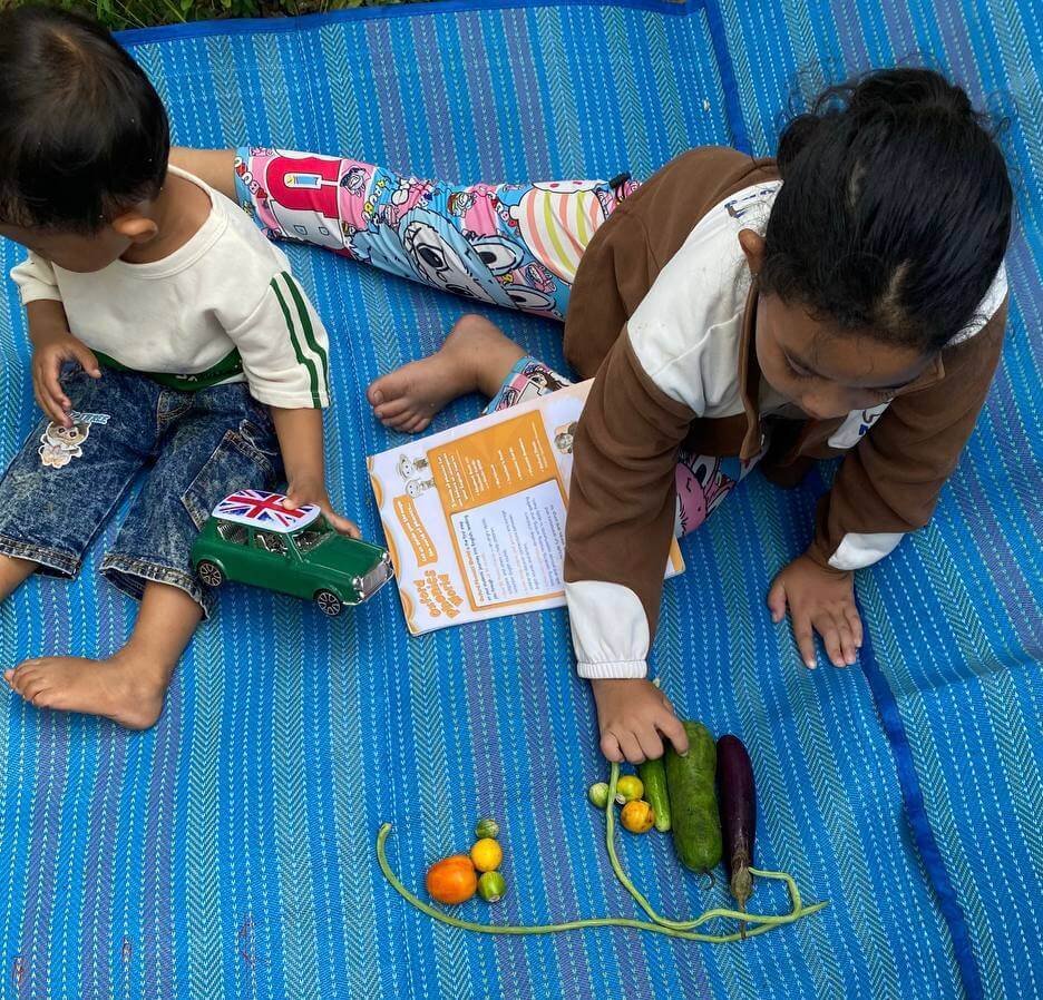 Two young children sitting on a blue outdoor mat with school books, looking closely at fresh vegetables including an eggplant, long beans, and small tomatoes.