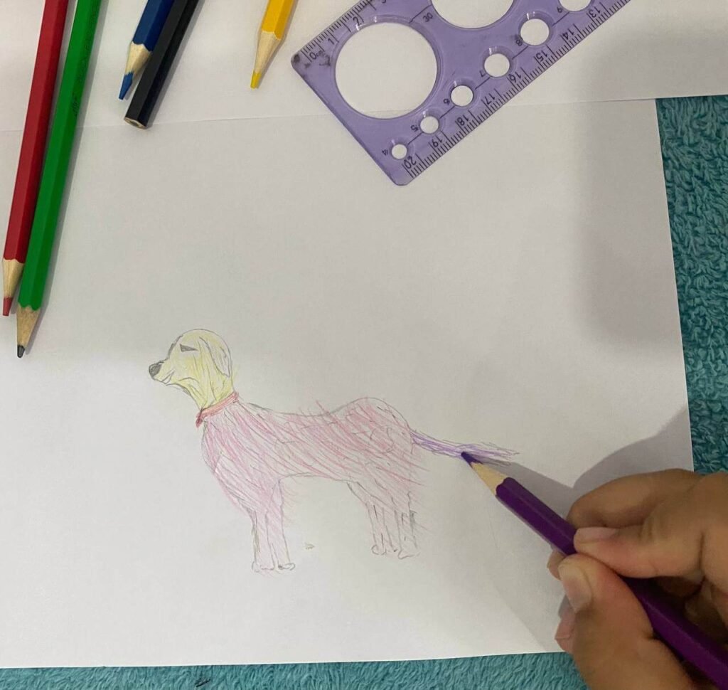 A finished drawing of a dog with a yellow head, pink body, and purple tail being colored by a child with a purple pencil.