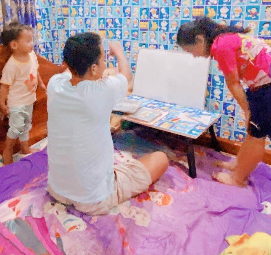 A father sitting on a bed next to a small folding table, giving his undivided attention to his two children without any screens nearby.